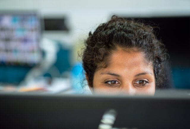 The top of a person's head is seen behind their monitor. They're looking at the monitor, not at the camera.